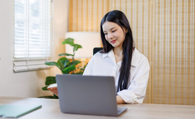 Adult Confident and smiling young Asian businesswoman work on laptop and use notebook from home office

