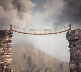 Suspended bridge connects two rocky cliffs in a misty mountain landscape during early morning