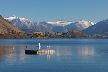 May 18, 2025: Stunning Lake Wanaka, South Island, New Zealand, with the majestic snow-capped Southern Alps reflecting in the clear blue water with swimming pier in the center. Tranquil alpine scenery. © Chonlatit