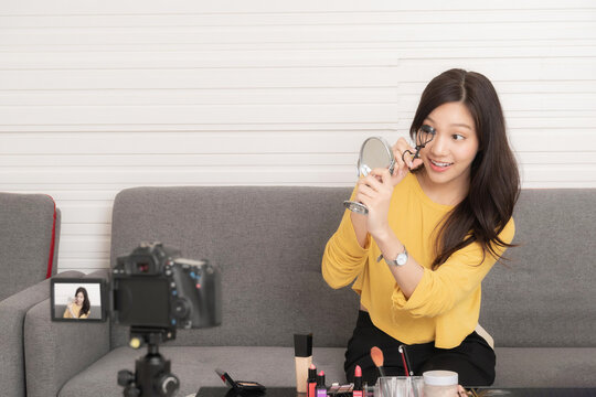 Young Asian beauty vlogger demonstrates eyelash curling using a mirror, while recording a makeup tutorial for her online audience.