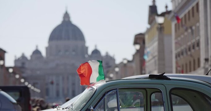 Cinematic Rome concept background. Italian flag waving in slow motion on vintage old timer car with St Peter Basilica at background viewed from Via della Conciliazione street