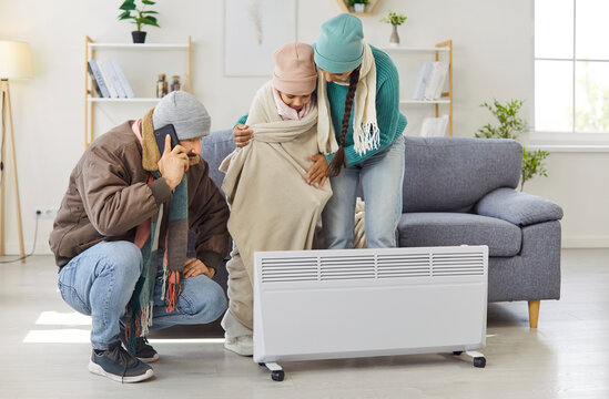 Phone call to emergency heating repair service, freezing family in sweaters and hats warming up by heater at home. Parents using electric radiator, hat and blanket to keep shivering child warm