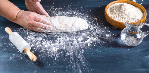 Female hands preparing fresh artisanal bread dough on a dark kitchen table. Baking ingredients flour, water, and a rolling pin are present