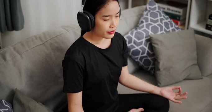Handheld high angle view shot, Asian woman practicing meditation and enjoying listening to music through wireless headphones on sofa in the living room at home.