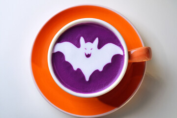 Top view of purple Ube latte in orange ceramic cup with Halloween-themed latte art on white background. This Ube latte features a bat halloween ghost design on milk foam. Generated image