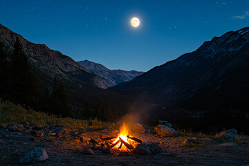 A campfire glowing in a mountain valley with the full moon overhead  