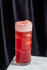 A cold, fizzy pink cocktail with a frothy top and ice rests on a white marble surface
