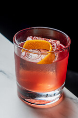 A vibrant red cocktail with ice and orange peel sits on a white marble surface