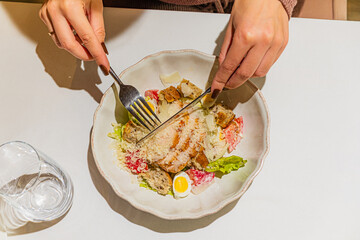 Person cuts chicken Caesar salad with utensils. Water glass nearby