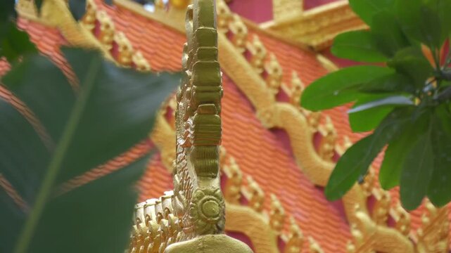 Majestic tiled Buddhist temple roof with golden intricate gable and serpent-like chofas stand in the surroundings of coconut palm and other trees crowns. Shot in motion