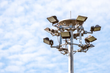 A cluster of powerful floodlights and spotlights on a tall metal pole, set against a bright, cloudy blue sky. This industrial fixture provides high-intensity stadium or area illumination.