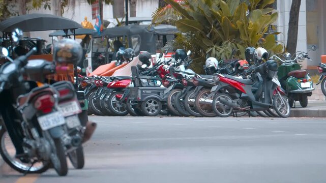 A scooter with a fully packed cart is driving near dense parking of scooters, motorcycles and mopeds on the road in Siem Reap, Cambodia