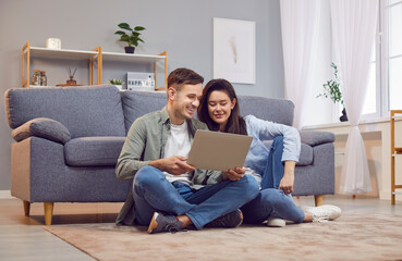 Young happy family couple sitting on floor in modern living room, looking at laptop screen. Smiling...