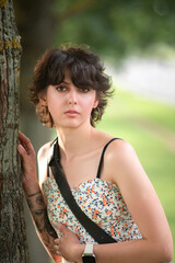Naklejka premium Young woman with short hair and floral dress leans against a tree, showcasing her tattoos and relaxed demeanor in a natural outdoor setting, capturing a moment of tranquility