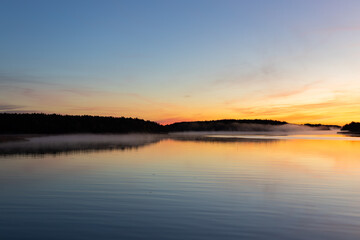 Golden sunrise over calm autumn sea with forest on the shore and gentle fog above the water, peaceful natural seascape and morning atmosphere