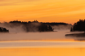 Golden sunrise over calm autumn sea with forest on the shore and gentle fog above the water, peaceful natural seascape and morning atmosphere