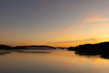 Golden sunrise over calm autumn sea with forest on the shore and gentle fog above the water, peaceful natural seascape and morning atmosphere