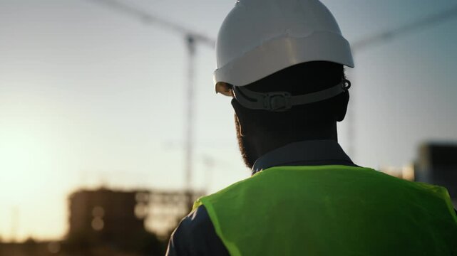 Builder walking at construction site, back view against sunset sky, slow motion. Middle-age male person correcting protective helmet on head, foreman or architect examining process of modern building