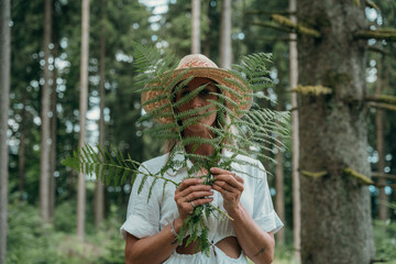 Green ferns cover part of a blonde woman's face in forest. She wears a straw hat. Eco and calm mood