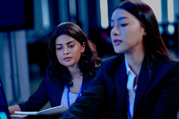 Intense late night focus: Two diverse female professionals collaborate closely, deeply engaged in reviewing critical project data displayed on a computer screen in a modern hub.