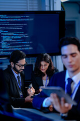 An AI research team brainstorms late night. Innovators discuss machine learning model architecture using a tablet while reviewing training data code displayed on a large screen.