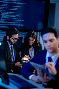 Focused diverse professionals collaborate late night in a modern tech office. Colleagues discuss project details using a tablet with stylus while code runs on a large screen.