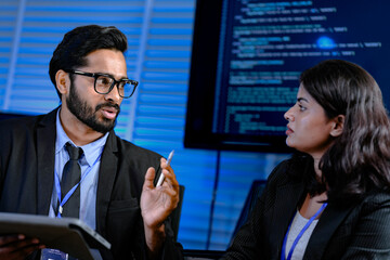 Focused late night strategy session. Male manager with glasses gestures explaining complex points to attentive female colleague in modern high tech command center office.