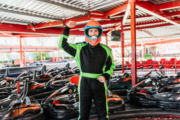 Young male karting racer in helmet and suit celebrating success, standing in a racing circuit grid...