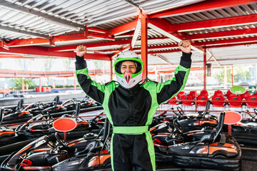 Obraz premium Young woman in a racing suit and helmet raising her arms, celebrating winning a go kart race at an outdoor track