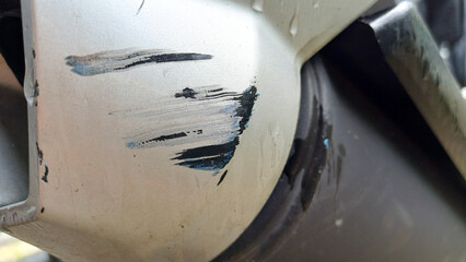 Close-up of deep, severe scratches, scuff marks with black and blue paint transfer on a light gray motorbike exhaust surface. Represents damage, accident, repair, texture, and insurance concept.