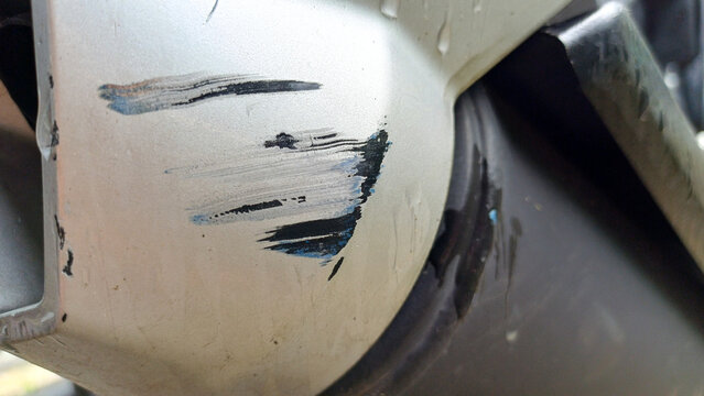 Close-up of deep, severe scratches, scuff marks with black and blue paint transfer on a light gray motorbike exhaust surface. Represents damage, accident, repair, texture, and insurance concept.