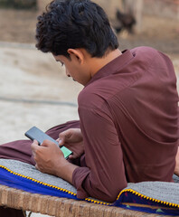 Young man sitting on traditional woven cot using smartphone outdoors in rural village setting