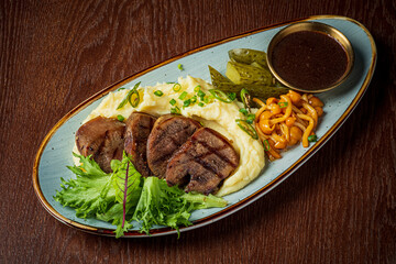 Grilled meat sits over fluffy mashed potatoes, accompanied by fresh greens and pickled vegetables