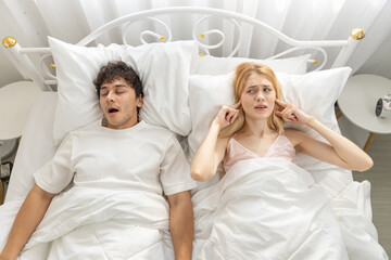 Frustrated women covering her ears while lying next to snoring man in bed, showing sleep disturbance and couple conflict in a bright white bedroom.
