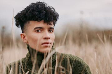 A close natural portrait of a young man in tall grass wearing a knit sweater, conveying authentic presence and reliable emotion with clear eyes and subtle expression in soft outdoor light.