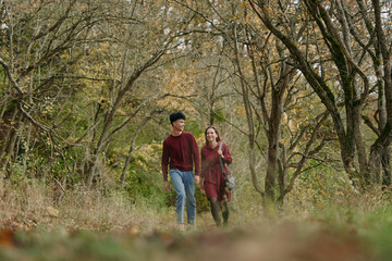 Fototapeta premium An authentic, candid couple walks through a sunlit autumn forest, showcasing real connection and casual style as leaves drift and light softens around them.