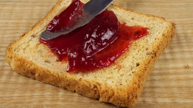 Redcurrant berry jam is spread with a knife onto a slice of multi-grain toast bread on a wooden board.