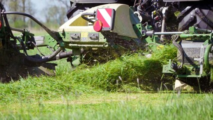 Close-up of modern agricultural machinery cutting and processing grass in green field during farming harvest season.