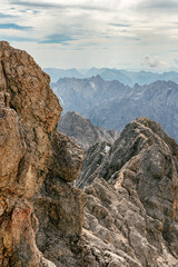Impressive view of the surrounding mountain peaks near Zugspitze in summer. Rugged rocks, snow patches, and distant valleys form a striking alpine landscape under a partly cloudy sky.