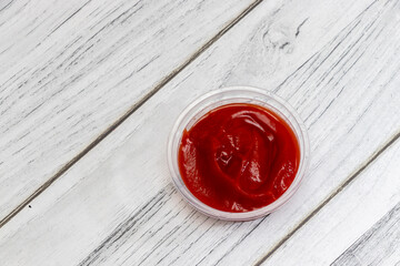 A close-up shot of ketchup in a clear container placed on a textured wooden surface, showcasing the rich red color, ideal for food photography or culinary presentations.