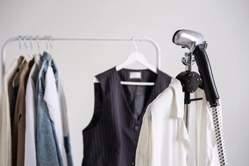 A clothes steamer stands next to a woman's fall wardrobe on a rail hanger. Clothes care.