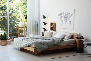 Cozy large bed with linens, furniture, houseplants and lamp near window in bedroom. Interior design