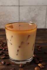 Tasty iced coffee with milk in glass and beans on wooden table against tiled wall, closeup