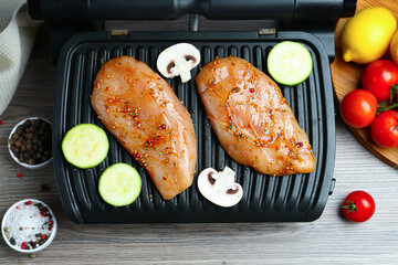 Electric grill with raw marinated chicken fillet, mushrooms, spices and products on wooden table, flat lay