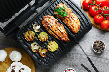 Electric grill with tasty chicken fillet, mushrooms, spices and vegetables on wooden table, flat lay