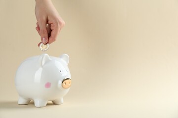 Saving money. Woman putting coins into piggy bank on beige background, closeup. Space for text