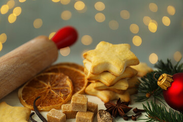 Christmas composition with cookies in shape of stars, spices, bauble and fir tree branches on light wooden table against grey background with blurred lights, closeup