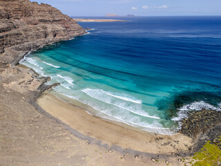 Aerial view of a secluded beach near Órzola, Lanzarote, framed by massive layered cliffs and turquoise Atlantic waters.
