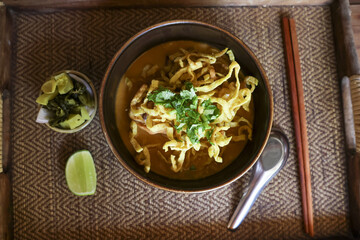 Savory Khao Soi, northern Thai noodle soup. An appetizing, comforting bowl of yellow curry with egg noodle, crispy topping, and lime on traditional wooden tray