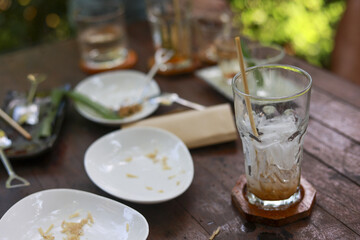 Messy wooden table at an outdoor cafe or restaurant after satisfying meal. scene shows an empty glass with ice, dirty plates, and leftover crumbs on tabletop